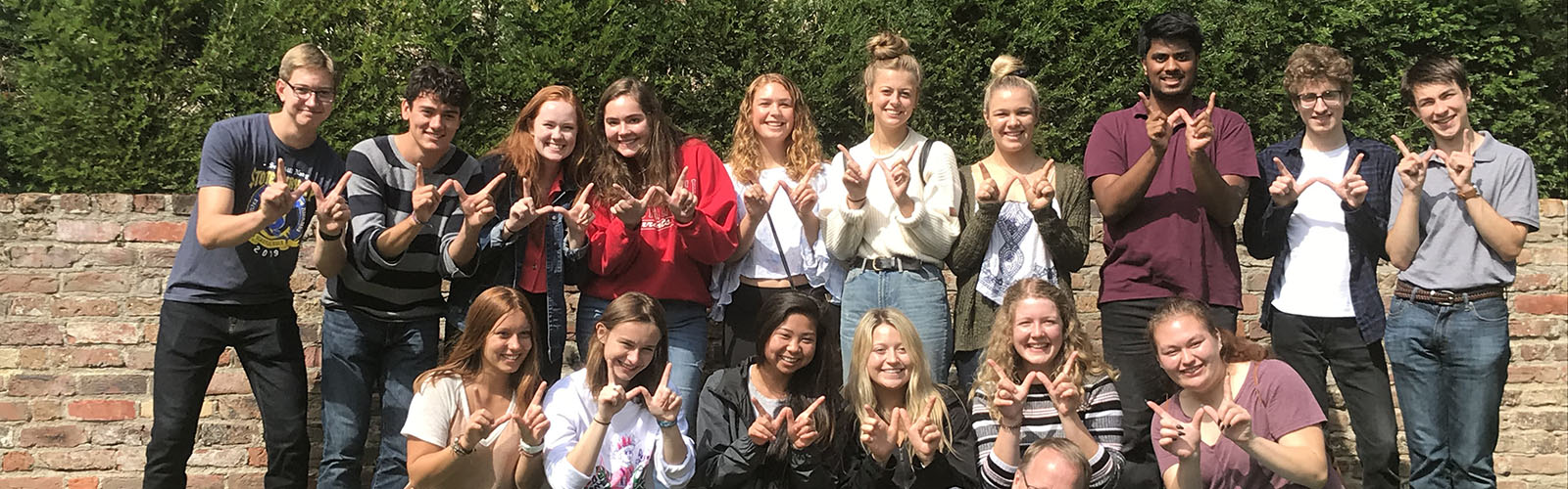 Fifteen students pose with the "Finger W" in front of a brick wall. 